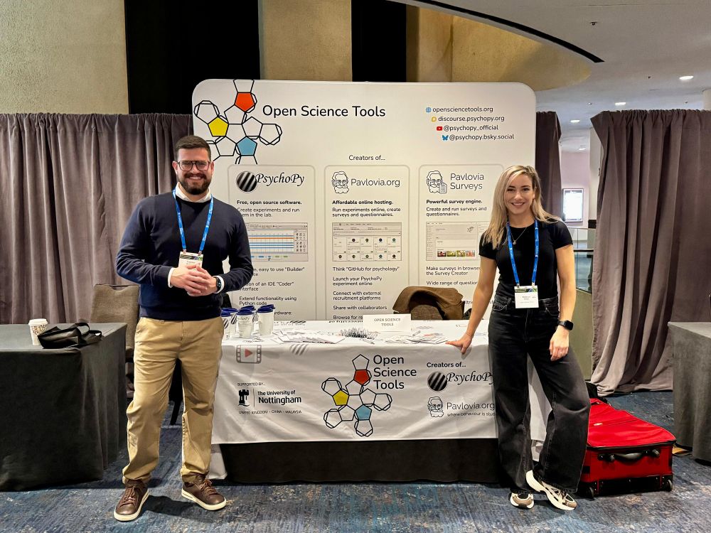 Two people standing Infront of a conference booth covered in merchandise. The booth reads "Open Science Tools" with products listed "psychopy", "Pavlovia", "Pavlovia surveys"