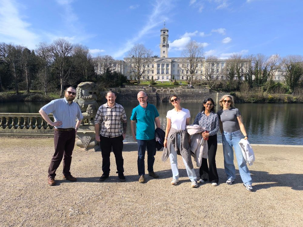 Six people stand happily in the sunshine Infront of the university of Nottingham Portland building, which can be seen across Highfields park lake.