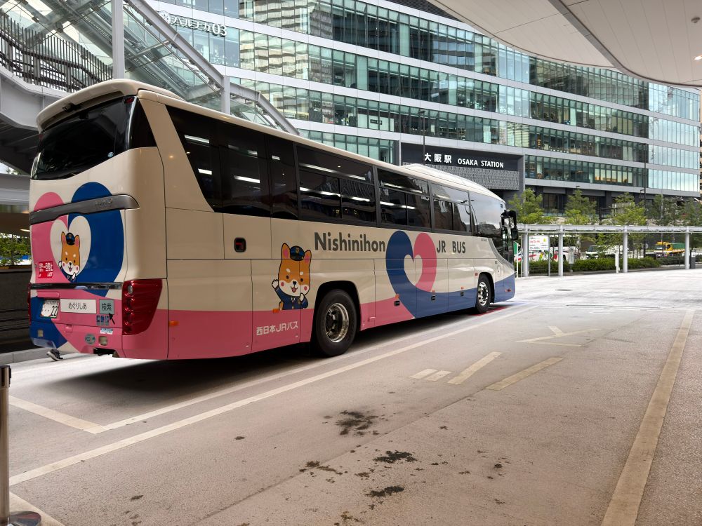 うめきたのバス乗り場に停車中のシャトルバスを斜め後ろから取った写真。白い観光バスタイプの車両で、そこにピンクと青色のラインでハート型をあしらった模様が描かれている。