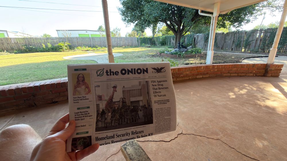 Image of The Onion newspaper in the foreground in someone’s hands with a green backyard in the background. There is a weird crocodile statue under a tree with two stone children sculptures riding it, and, baby, you know that’s what got this person to rent this house. 