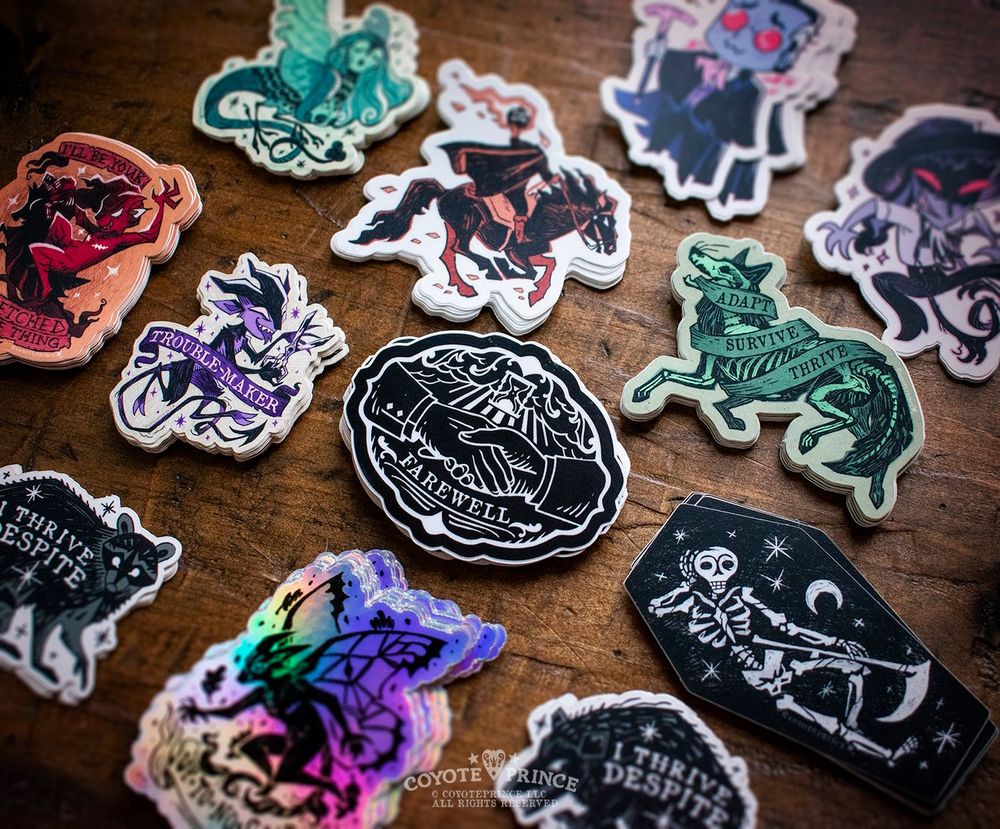 A photo of colorful Gothic stickers neatly aligned on a wood table. The angle is skewed. The stickers show a variety of fairies, momento mori, and North American animals.