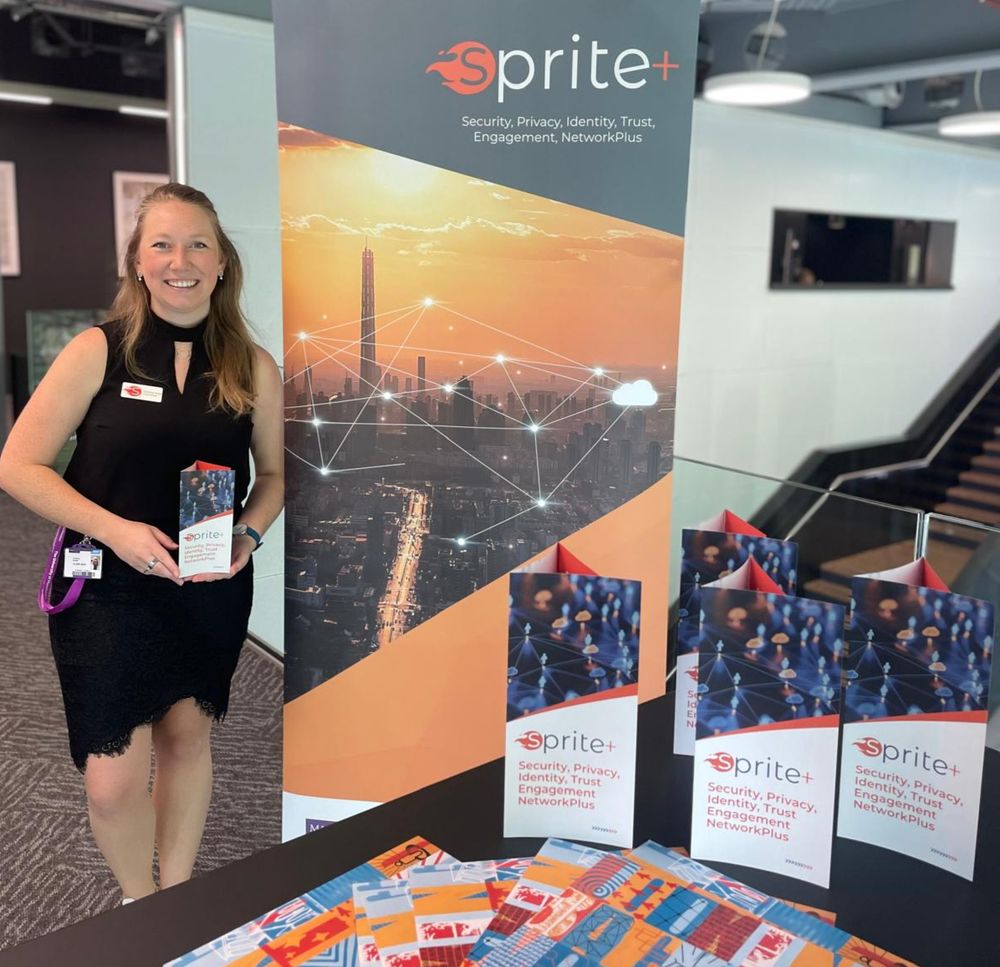 Courtney, a fair-haired woman in a black dress, stands next to a banner which reads 'SPRITE+ Security, Privacy, Identity, Trust, Engagement, NetworkPlus', holding a SPRITE+ information leaflet. 