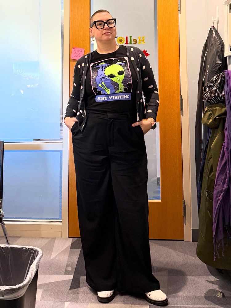 A white non-binary person wearing a tshirt that has an alien and a spaceship abducting a cow that says “just visiting,” with black pants, a polka dot cardigan, and white shoes
