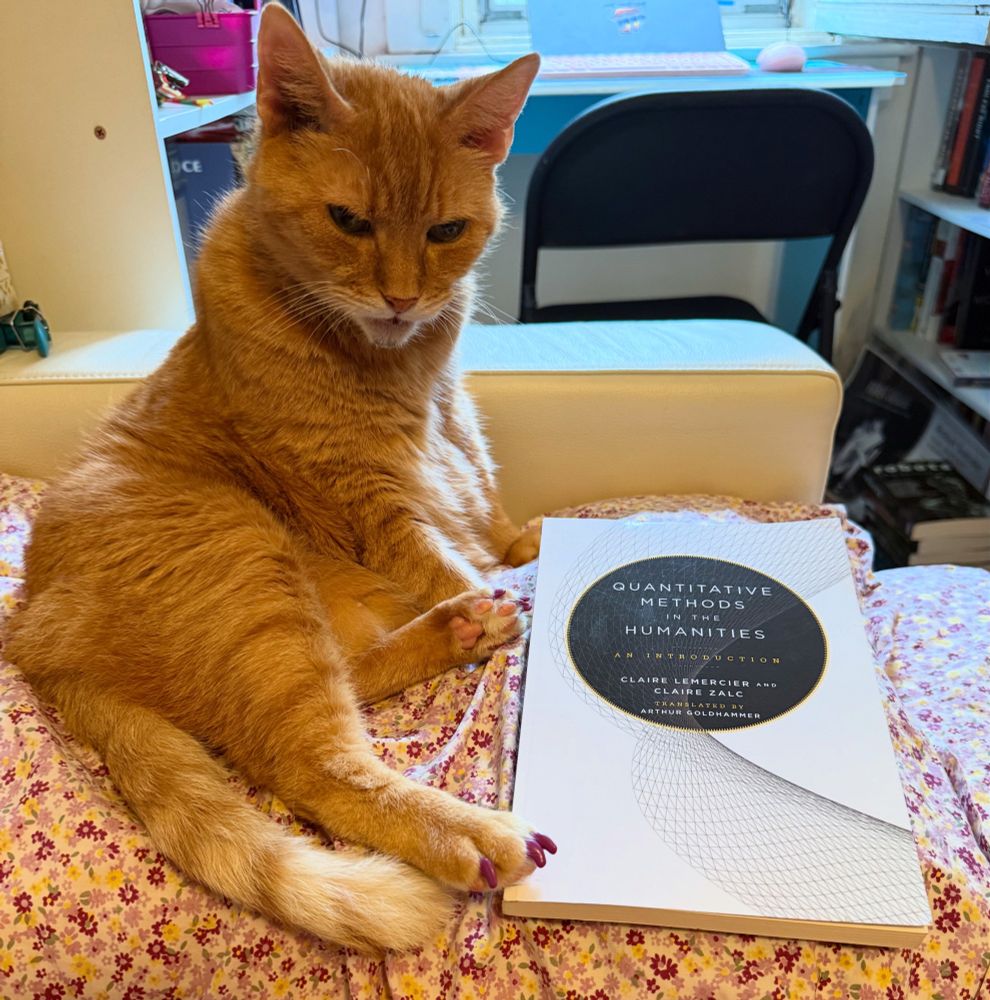 An orange cat sitting on her side, looking grumpily at the camera. Beside her is a copy of Quantitative Methods in the Humanities by Claire Lemercier and Claire Zalc (translated by Arthur Goldhammer). 