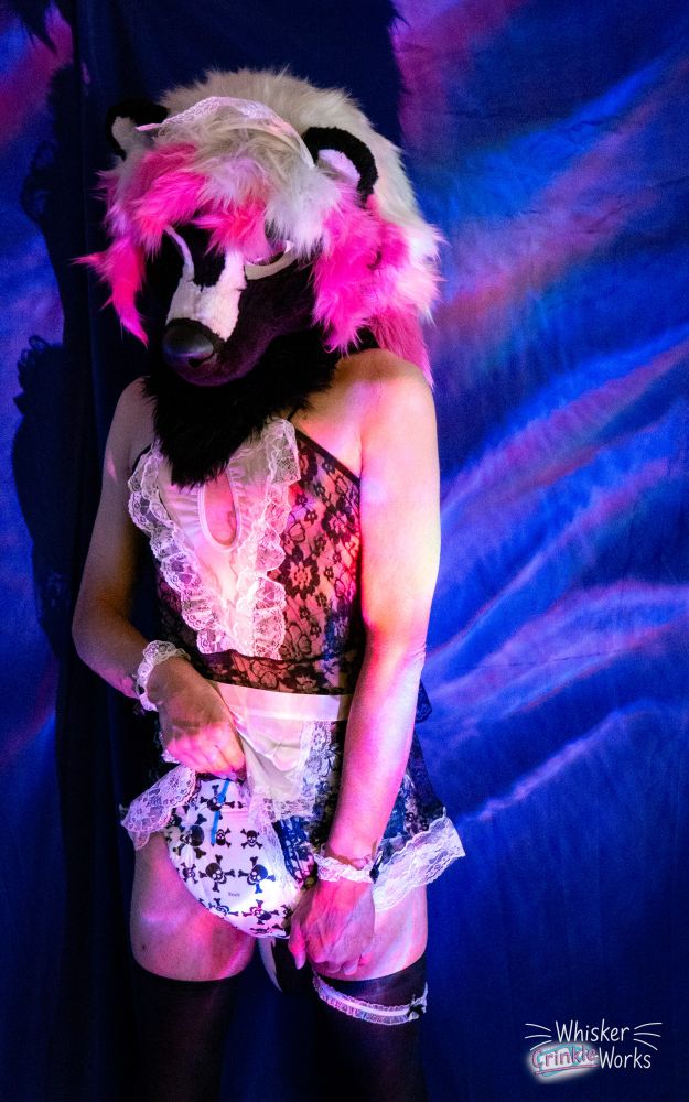 A black and white furred skunk partial fursuiter, with pink and silver hair, is looking down and off to the side, eyeing the camera in a teasing way. She is wearing a see through maid outfit with black leggings. She is lifting one side of the skirt up while continuing to hold the other side down, giving a good look at the diaper underneath- a discontinued diaper, Rearz Rebels, to match her fur and outfit colors and to tease about being a skunk and having to deal with her diaper scent.