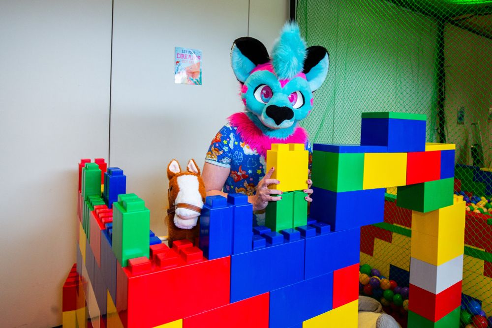 A blue, pink and black possum fursuiter in her head and a blue onesie, playing with giant blocks in a playroom during the previous Babyfur Con. The blocks are stacked like a small castle wall, with an empty doorway small enough to have to crawl through. There is also a plush horse next to her and a ballpit as well on her other side.