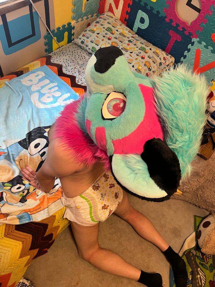 A partial fursuiter in a fursuit head. The fursuit head is a pink and blue and black furred animal with a blue mohawk and pink and blue eyes. Their nose is black. They are staring almost upward as they hold onto a bed sitting on the floor below them while kneeling on the floor. They are wearing a thick crinklz diaper as well. The room around them, including the bed's blankets are very kid/baby designed.