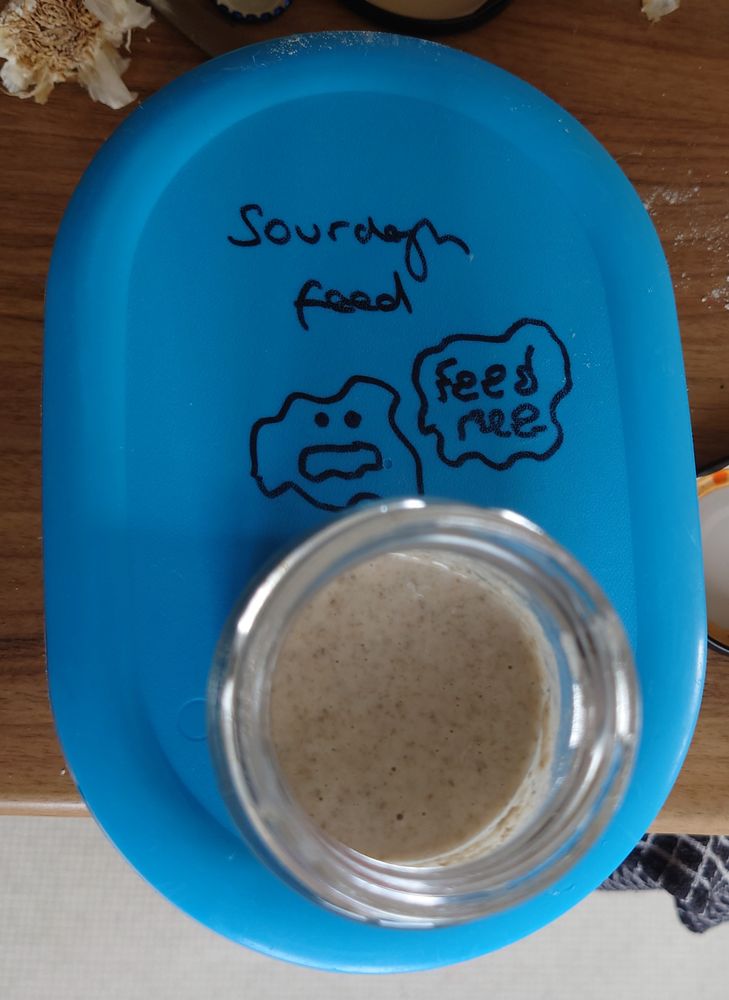 A small glass jar of sourdough starter sat on a blue lidded plastic tub. The lid is labelled "sourdough feed" and has a drawing of a blob with eyes on it, saying "feed me"