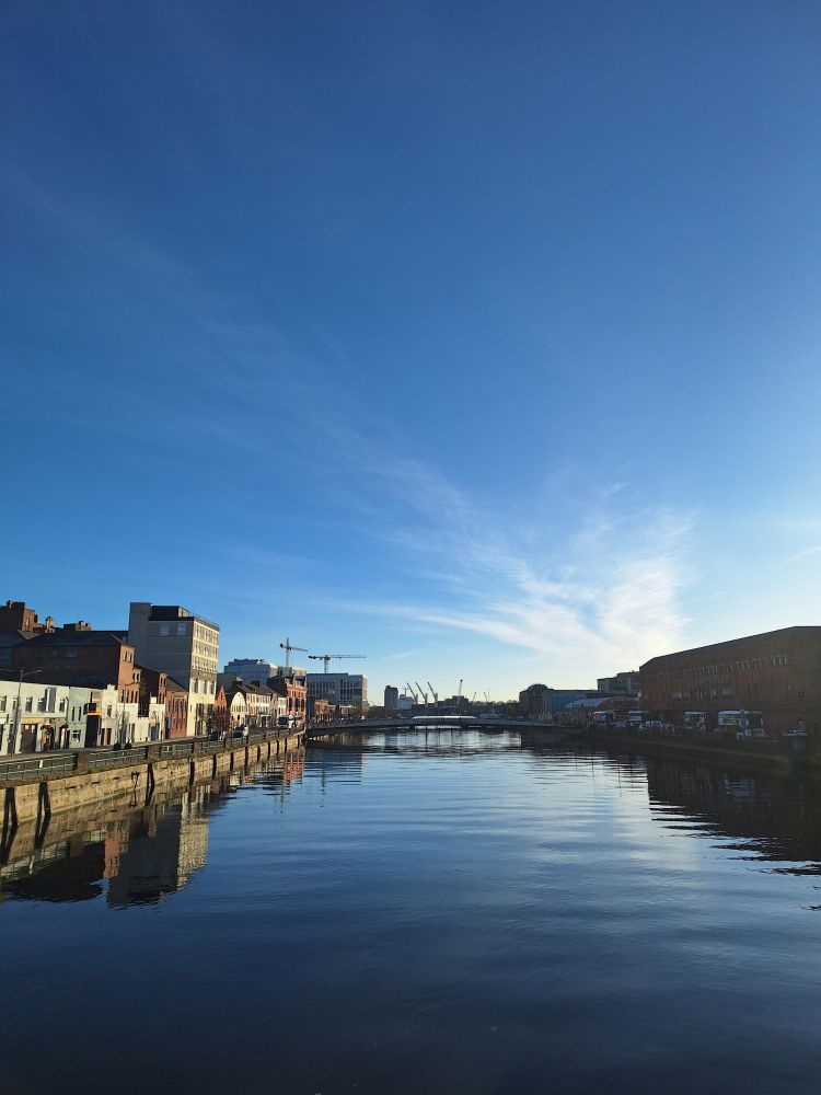 A view of a river running through a city, almost cloudless blue sky reflected in the water, bright early morning sunshine shines on the buildings on the left bank while the buildings on the right are in shadow