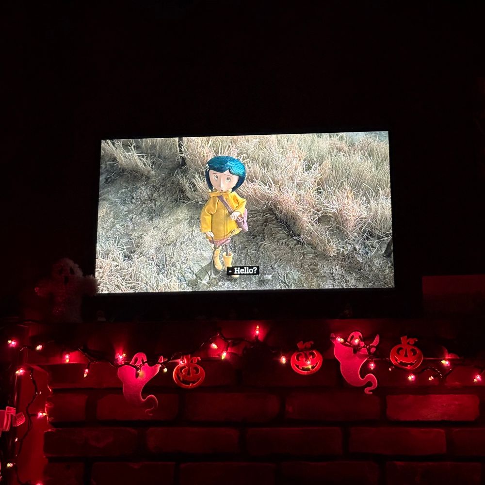 A photo of a tv with the movie Coraline. She has blue hair and a yellow rain coat and yellow boots. The tv is on top of a mantle with purple lights with ghost and pumpkin decor. 