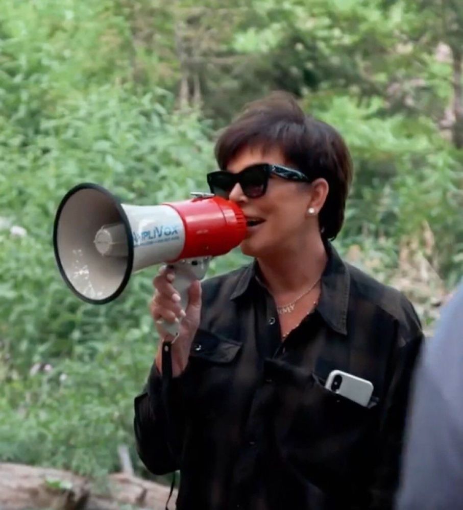 Kris Jenner holding a megaphone to her mouth. She is wearing black sunglasses, a black long sleeve blouse, and white stud earrings. She has a white iPhone tucked into her breast pocket. The background are green bushes. 