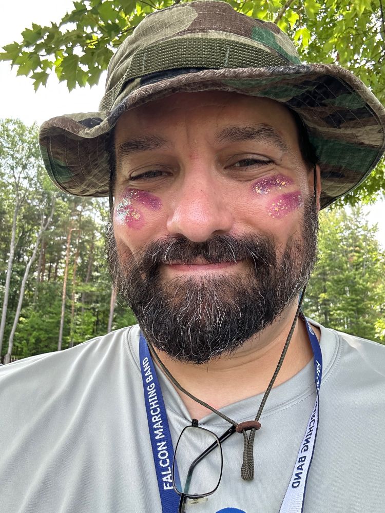 Bearded adult male wearing a camouflage hat with four streaks of glittery makeup on his cheeks. 