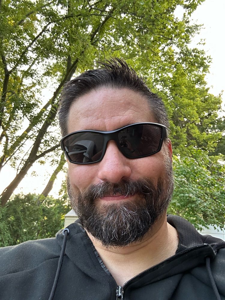 Beardy white guy in sunglasses smiles slightly in front of some leafy green trees.