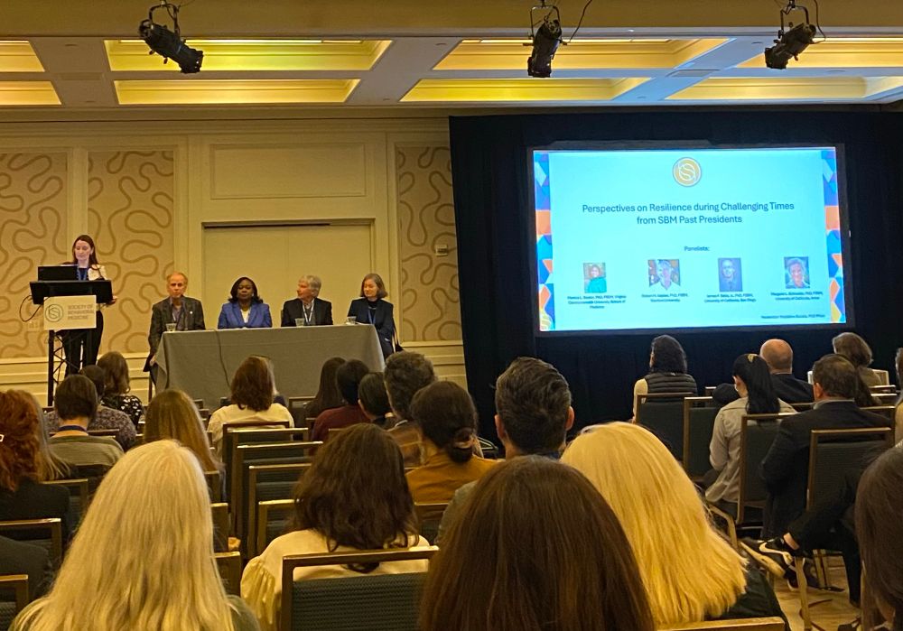 Four panelists (2 men and 2 women) and a speaker (woman) in front of an audience with a large slide displayed, “Perspectives in Resilience during Challenging Times from SBM Past Presidents”