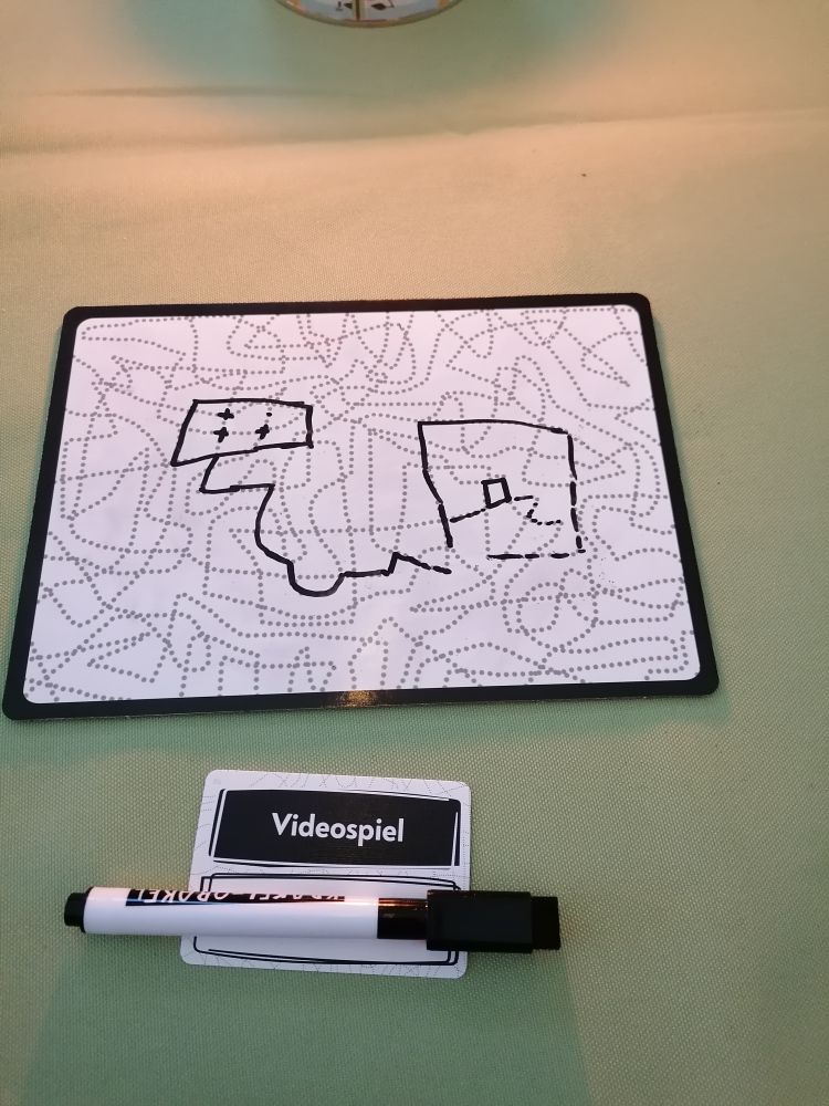 Eine weitere Krakel Orakel Tafel, auf der mit Strichen eine Art Videospiel-Controller mit Kabel und ein Bildschirm mit einer Röhre auf einem Weg dargestellt. Als Begriff auf dem Kärtchen steht "Videospiel".