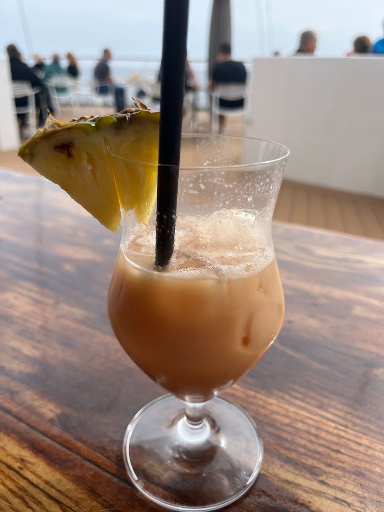 Ein Cocktailglas mit einer orangefarbenen Flüssigkeit, einem Strohhalm und einem Stück Ananas. Das Glas steht auf einem lackierten Holztisch