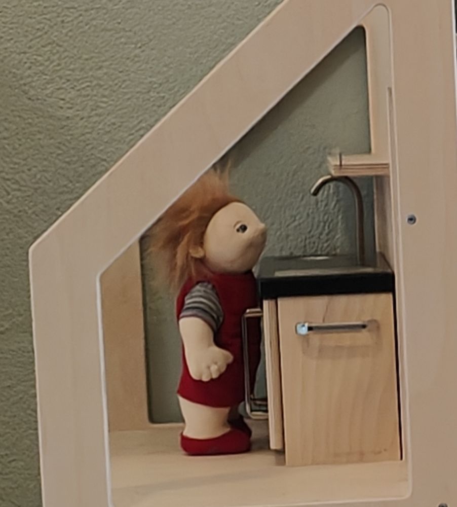 Stoffen popje wat bij het aanrecht staat in een houten poppenhuis. 