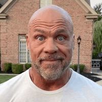 a bald man with a beard is standing in front of a brick house and making a funny face .