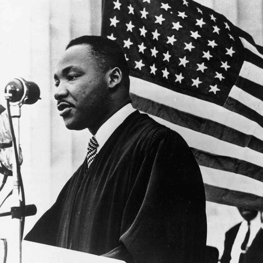Black and white image of MLK standing at a microphone, with a U.S. flag behind him.