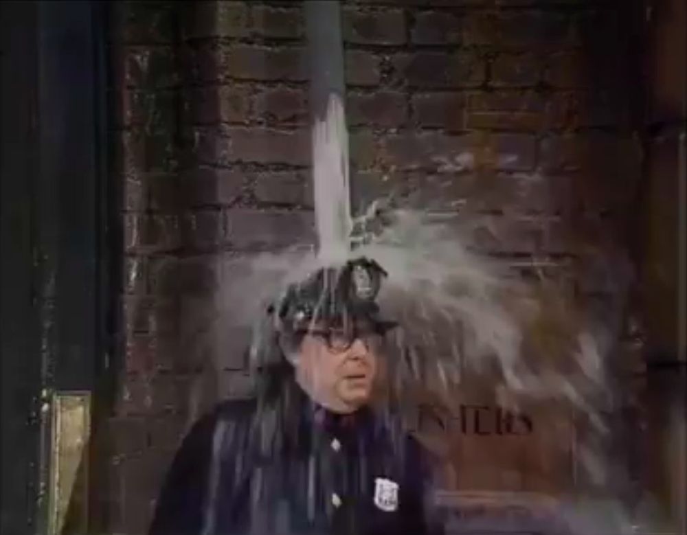 Eric Morecambe dressed as a policeman getting drenched by water from a down pipe.