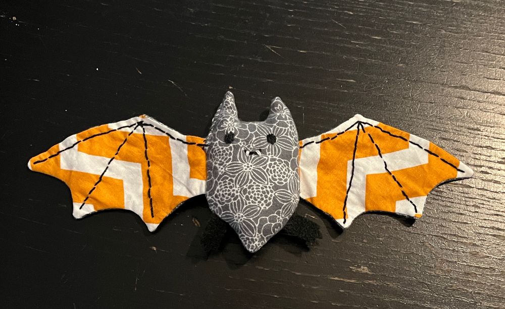 Simple shaped bat plushie made out of gray and orange fabric. It has a little face embroidered on it and stitching on the wings. 