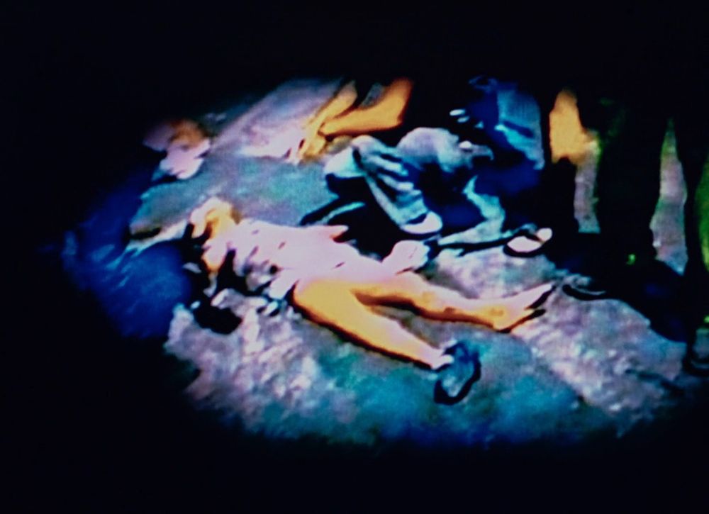 Image très alterné, une tache entourée de noir, couleurs primaires exacerbées, presque baveuses, tendant vers le bleu pour le sol et les vêtements, vers le jaune ocre pour la peau humaine, d'une personne étendue inanimée, que d'autres personnes viennent voir