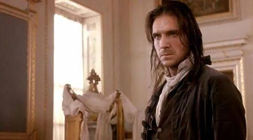 Ralph Fiennes no filme O Morro dos Ventos Uivantes. Ele está de cabelo comprido preso, casaco marrom escuro e um lenço branco no pescoço e olha bravo para algo. Está numa sala branca com detalhes de molduras douradas e uma janela ao fundo.