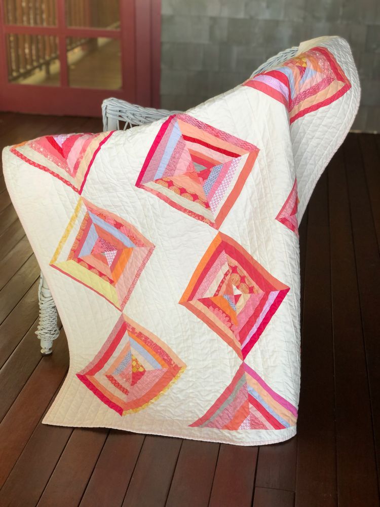 Quilt draped on chair. Diamond set blocks using scrap fabrics in tones of peach, pink, yellow and orange on a cream background.