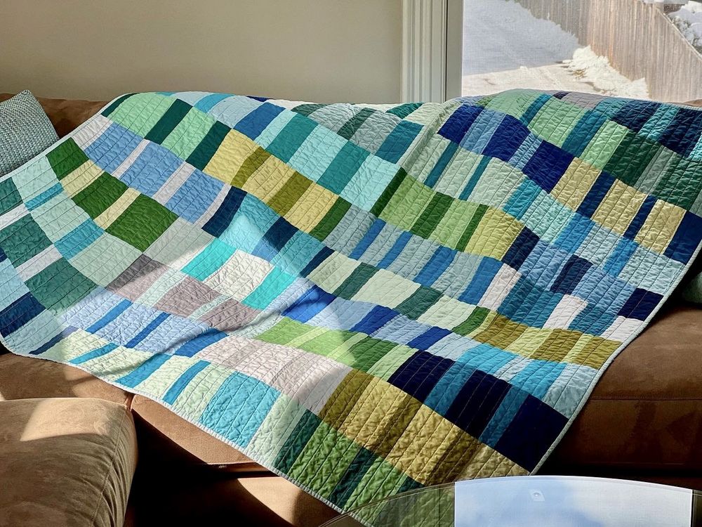 Quilt with vertical bars of color laying on sunny couch. Colors are shades of blue, green, yellow, white and grey. 