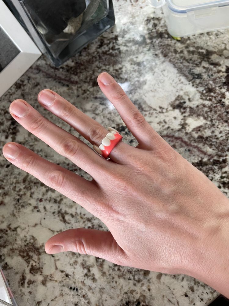 photo of the author's hand modeling a plastic ring. The ring is pink and white and shaped like vampire teeth.