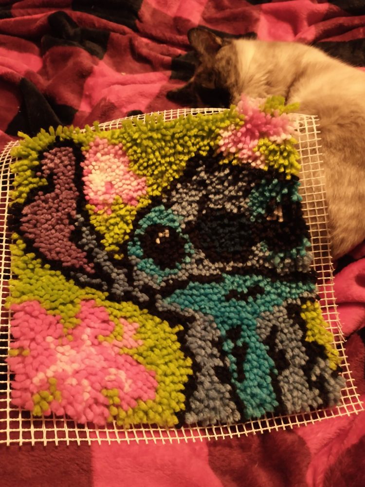 Stitch latch hook rug completed laying on top of a Siamese cat sleeping on a red and black blanket