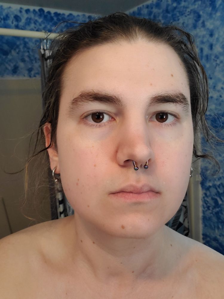 a girl (me) dark hair with blonde streaks tied back in a bun. curved barbell piercings in septum and lobes; stainless in lobes, rainbow anodized in septum. no shirt but not lewd; the tops of my shoulders are showing. there is a shower behind me