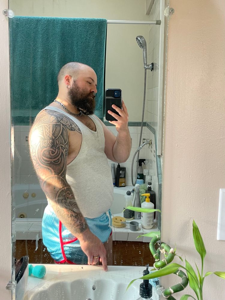 Bald bearded guy poses to the side for a mirror selfie to show off developing thickness