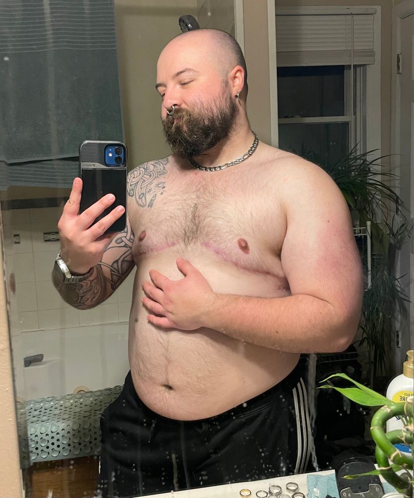 thicc white transmasc poses in front of a bathroom mirror. He has a shaved head and rough beard.