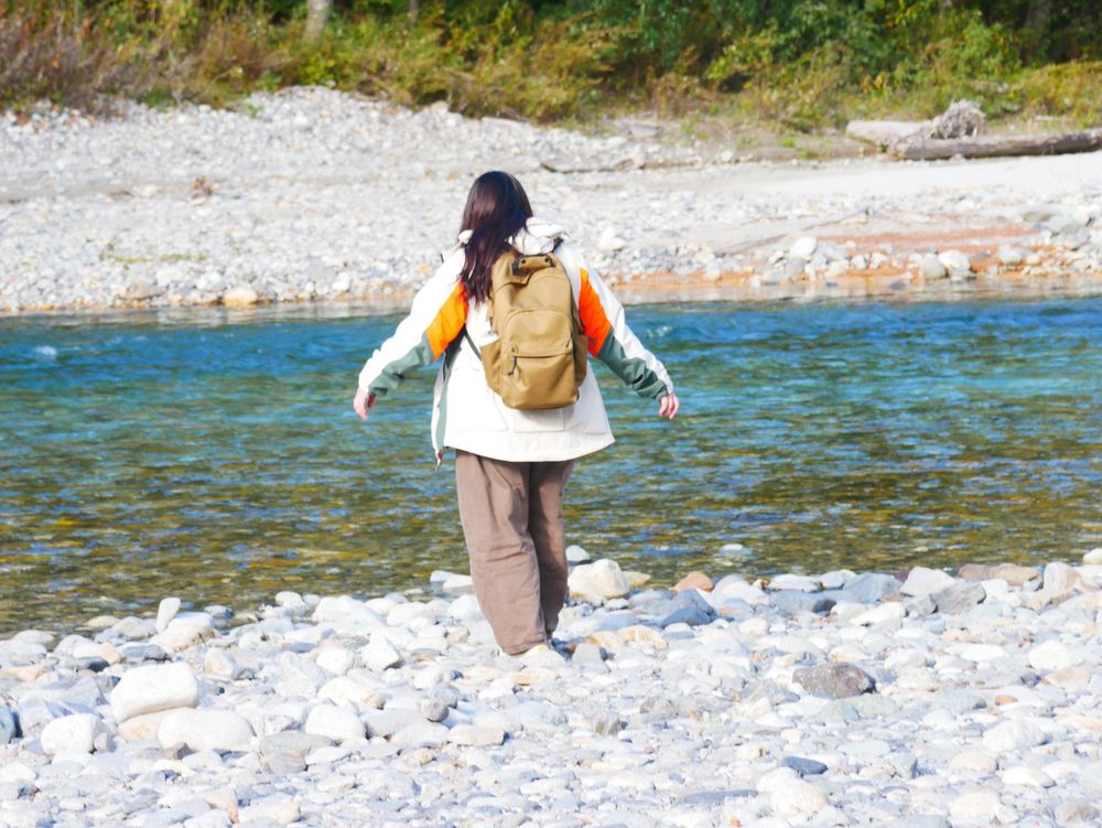 青い清流へ吸い寄せられるように足取り軽く河原を進む人物の後ろ姿。濃いベージュのリュックを背負い、白とオレンジ、グリーンの切り替えの入ったアウターを羽織っている。
