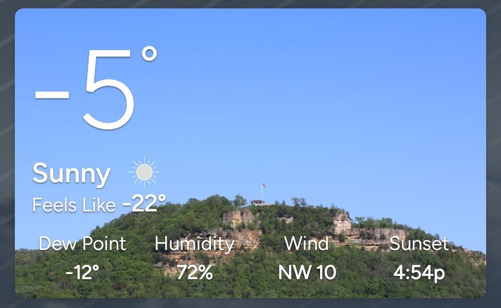A screenshot of current weather conditions showing a temperature of -5°F with a Feels Like temp of -22°F. The background is a bluff top with a flag and structure in the distance.