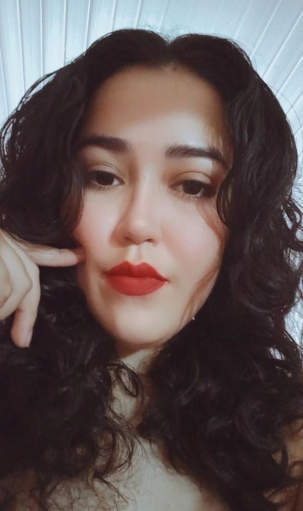 A foto mostra uma pessoa em close, do peito para cima, com os cabelos escuros, soltos e ondulados, caindo ao redor do rosto. Ela usa batom vermelho intenso e maquiagem leve nos olhos e sobrancelhas. O rosto está levemente apoiado na mão esquerda, com o dedo indicador tocando a bochecha. O fundo é claro, com linhas diagonais que lembram uma superfície de tecido ou forro.