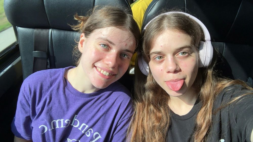 Selfie of Tzipporah and Yocheved in bus seats. Tzipporah is wearing light pink headphones and a black shirt and sticking their tongue out. Yocheved is wearing a purple shirt and smiling.