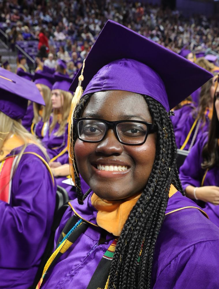 Photograph of Ellie smiling at the camera from her aisle seat at her college graduation. Everyone wears purple caps and gowns. 