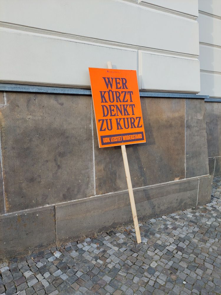 Ein Schild mit der Aufschrift "wer kürzt denkt zu kurz" lehnt an einer Wand. 