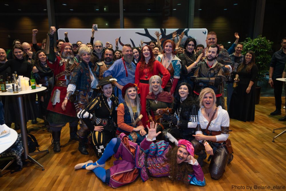 A large group of The Witcher in Concert attendees, cosplayers, and performers pose together for the camera. Many raise their hands in celebration and smile. At the bottom right corner text reads, "Photo by @‌elarie_elarie".