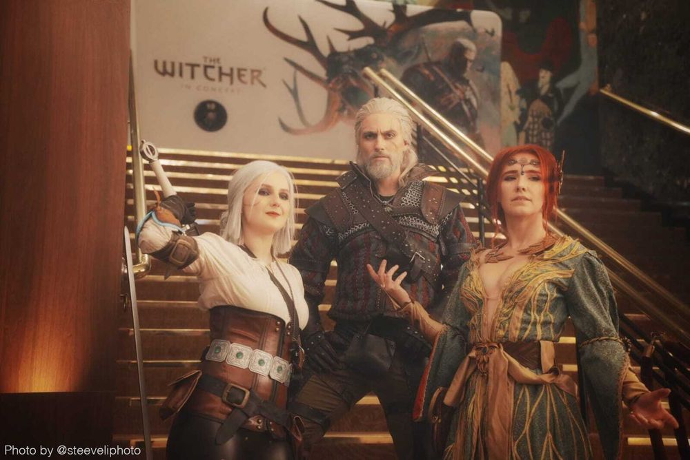 Three cosplayers dressed as Ciri, Geralt, and Triss pose at the foot of the stairs. Behind them is a large banner that reads, "The Witcher in Concert".