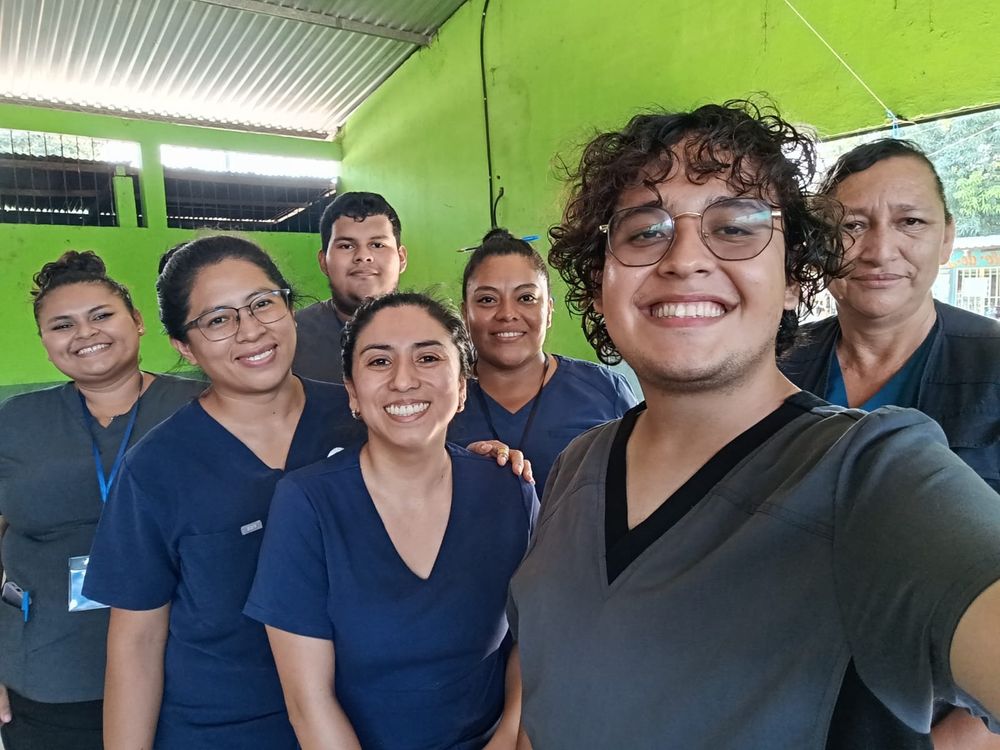 7 team people all with dark hair and wearing scrubs stand in a room that is painted bright green