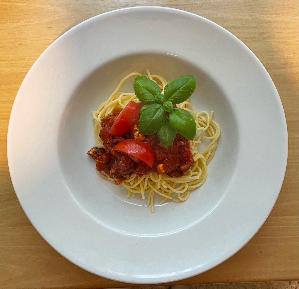 Zu sehen ist ein Teller mit Spaghetti, Tofu-Bolognese Sauce und Tomaten , garniert mit einem Basilikumblatt