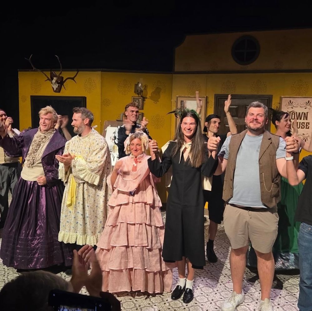 Screenshot from Rosie’s IG of Rhys participating in a Fringe show wearing a big shiny purple dress with a cream collar and panel running down the front. About a dozen participants are on stage as well some in costume and some in street clothes 
