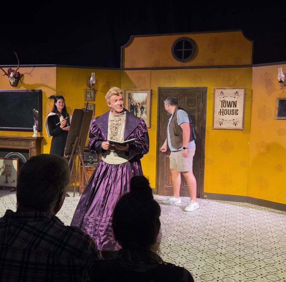 Screenshot from Rosie’s IG of Rhys participating in a Fringe show wearing a big shiny purple dress with a cream collar and panel running down the front. He is holding a book? There are some people behind him, somebody looks like they’re maybe painting?