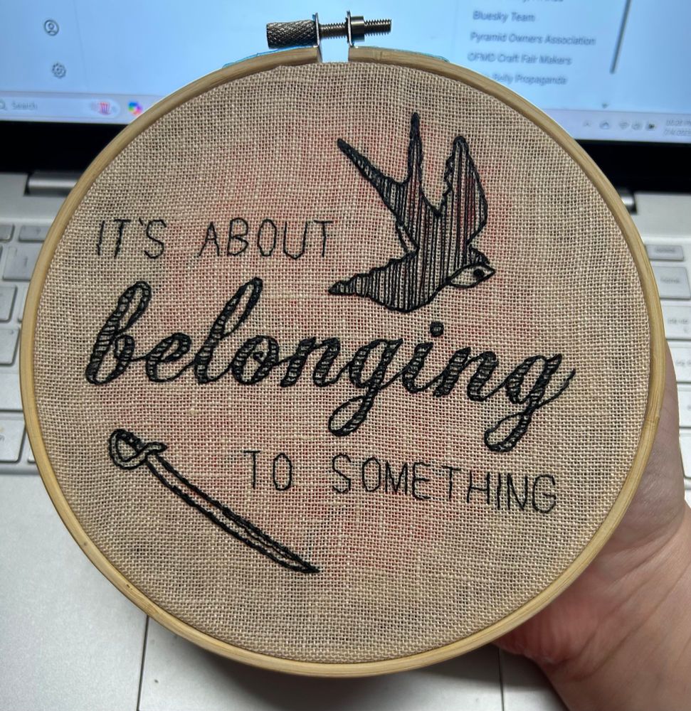 Photo of an embroidery that says IT’S ABOUT BELONGING TO SOMETHING in black thread, there is also a swallow and a sword in the piece, and the word belonging is in a decorative script while the rest of the words are sans serif letters. There is a circle of orange fabric juuuuuuust visible through the project’s fabric, at the back. Woops!