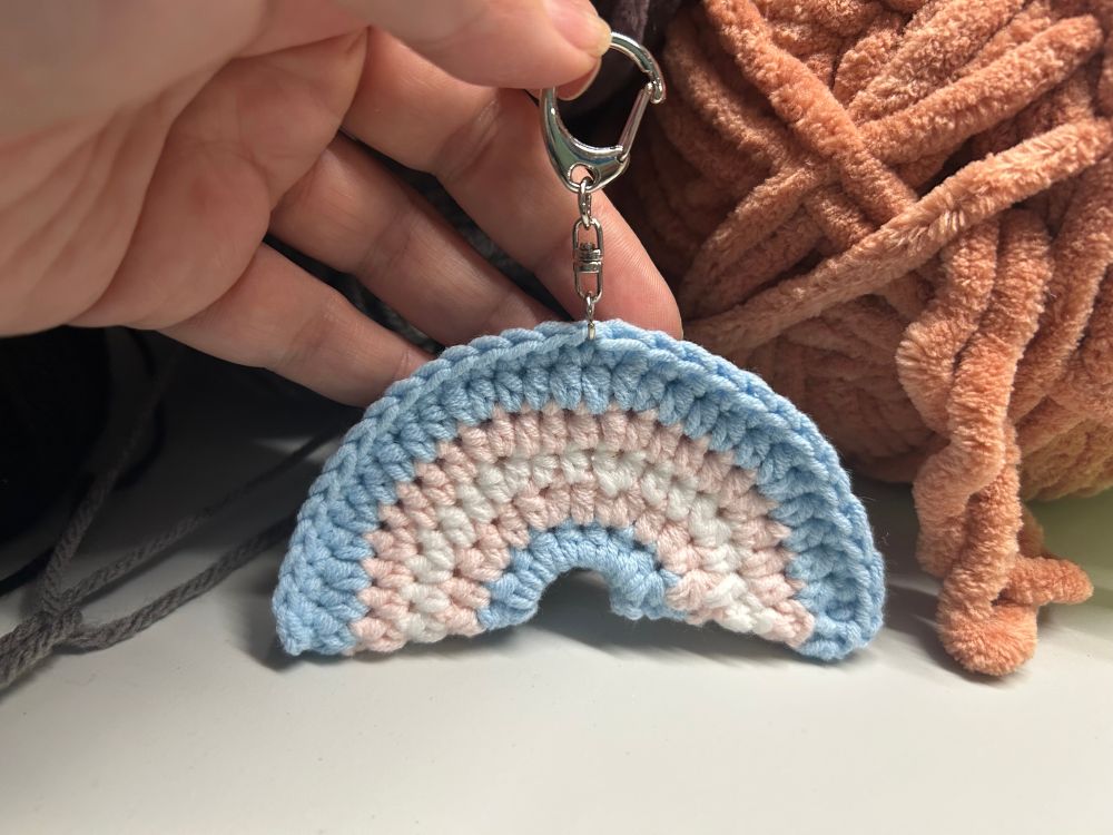 Photo of a small crocheted rainbow in the trans pride flag colors, blue, pink, white, pink, and blue. It is held up by a keychain attached to the top of it