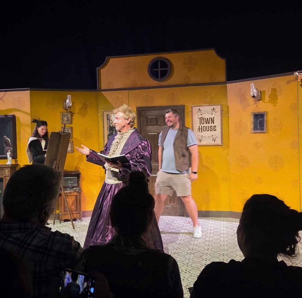 Screenshot from Rosie’s IG of Rhys participating in a Fringe show wearing a big shiny purple dress with a cream collar and panel running down the front. He is holding a book? There are some people behind him, somebody looks like they’re maybe painting and a guy in shorts, t shirt, and vest is laughing