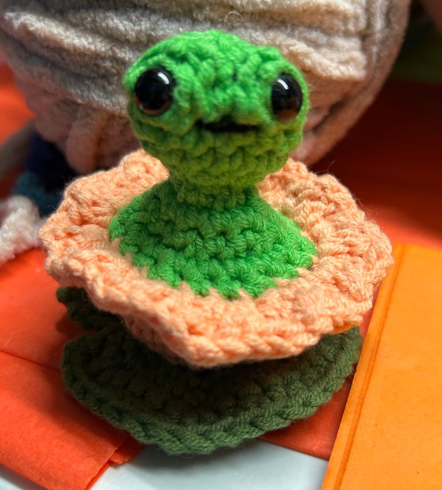 Photo of a crocheted green frog that looks kind of like an alien sitting in a peach flower on top of a dark green lily pad. The frog is peeking way up out of the flower.